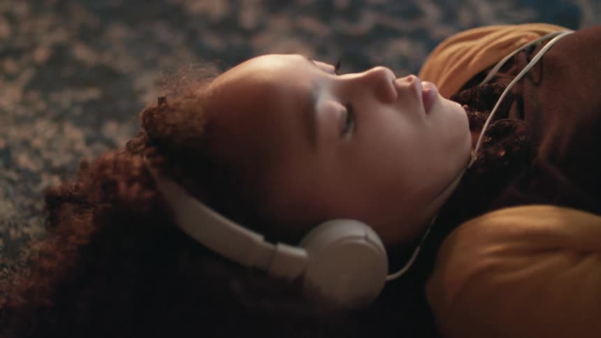 A little girl laying on the floor on her back wearing a pair of headphones while scrolling on a smart phone