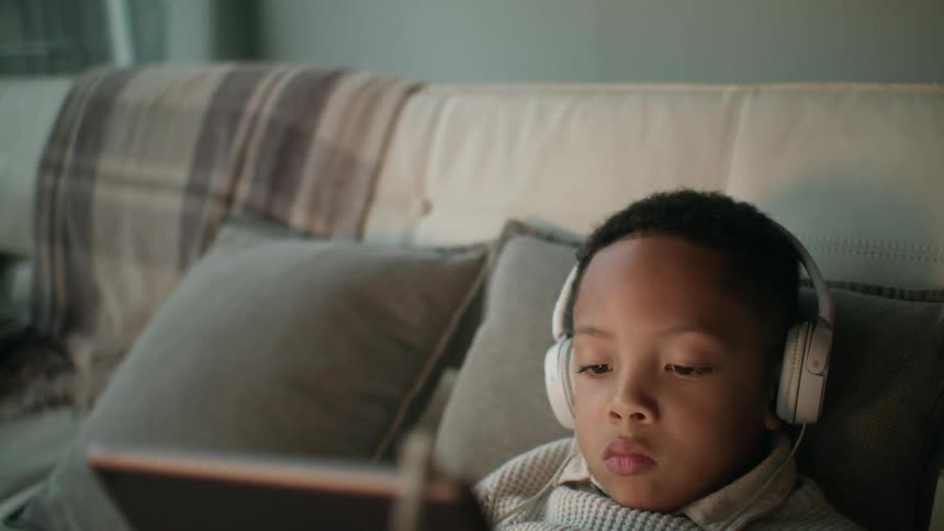 A young boy sitting on a sofa is wearing headphones and doing something on a tablet