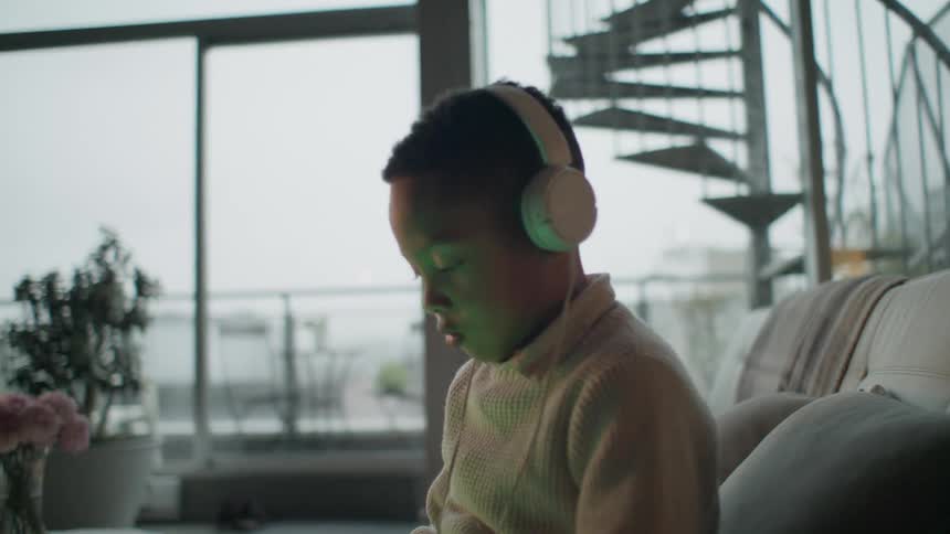 A boy in a room sitting on a couch looking at a tablet with headphones