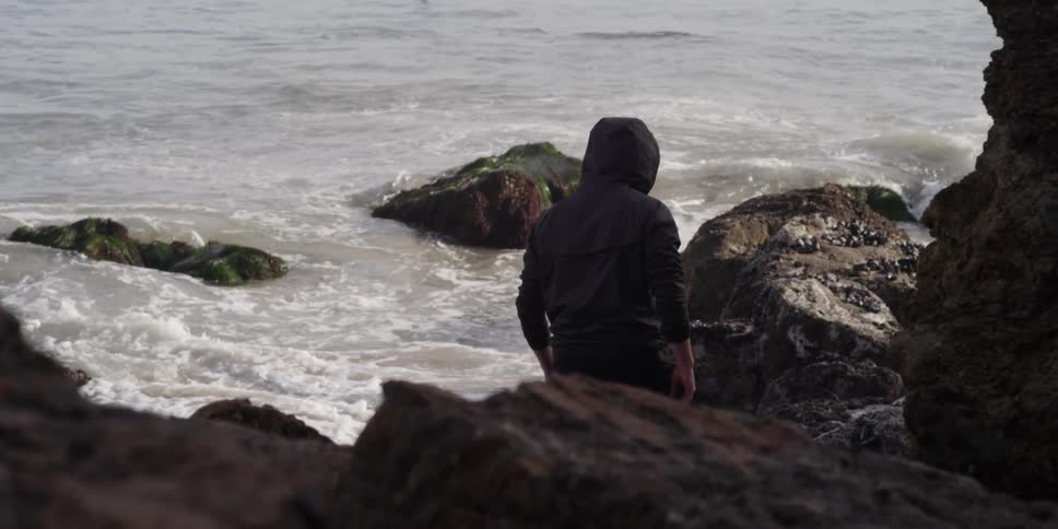 A man walks along a rocky shore while another is out in the water