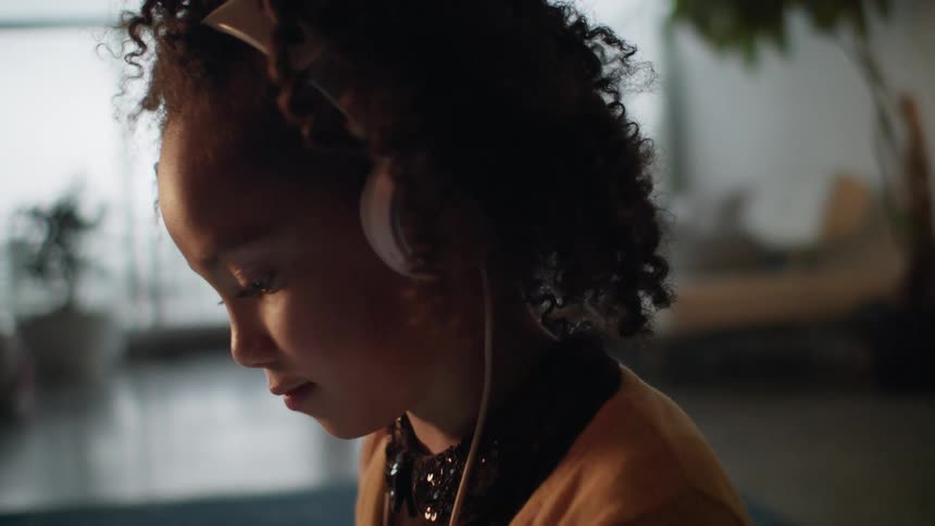 A girl uses headphones to listen to a tablet while sitting on a rug in the living room of a home during the day