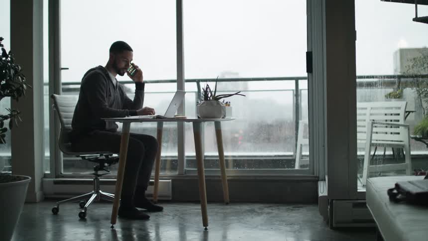 A man uses his smartphone while sitting at his desk at his downtown apartment on a rainy day