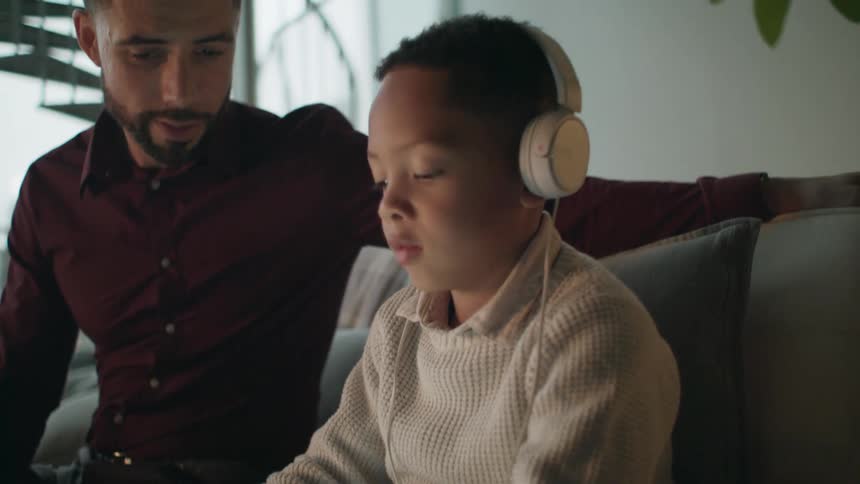 A father watching his young son play on a tablet while sitting on a living room sofa