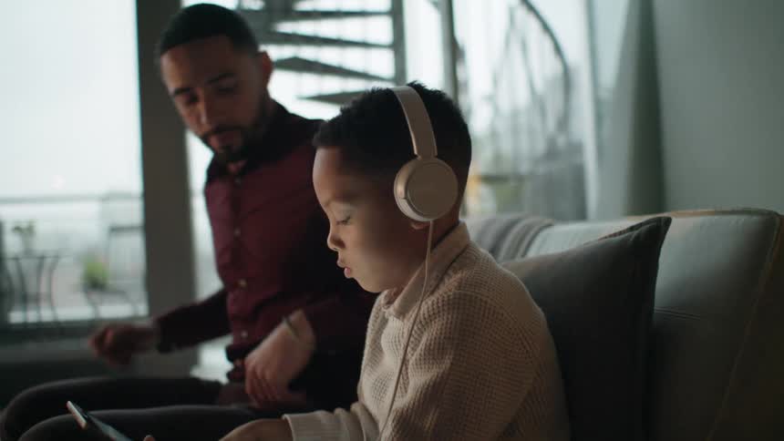 A boy sitting on a couch with headphones looking at a tablet as a man sits and talks to him