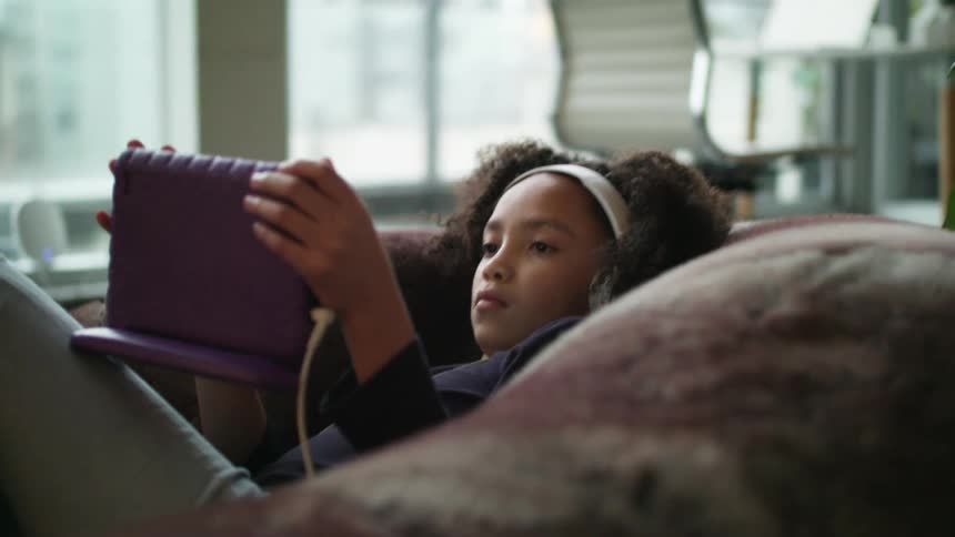 A girl laying on a couch playing with a tablet with a chair and a window in the background