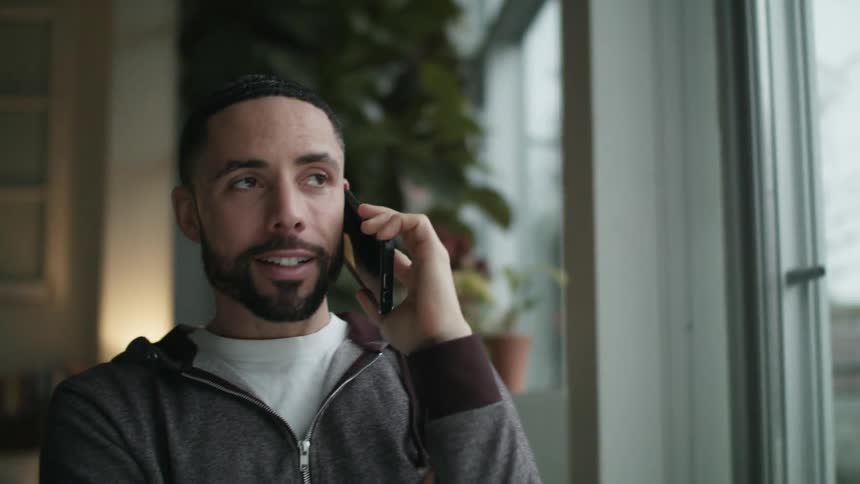 A man in a room talking with a smart phone next to a window with a lamp and a wall in the background
