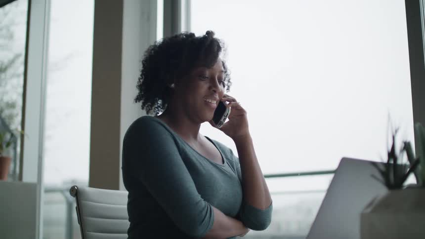 A woman in a room sitting on a chair talking on a smartphone next to a laptop