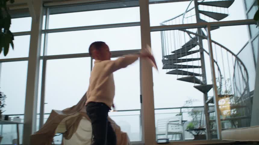 A boy plays with a paper airplane in the living room near a play tent and large windows and doors