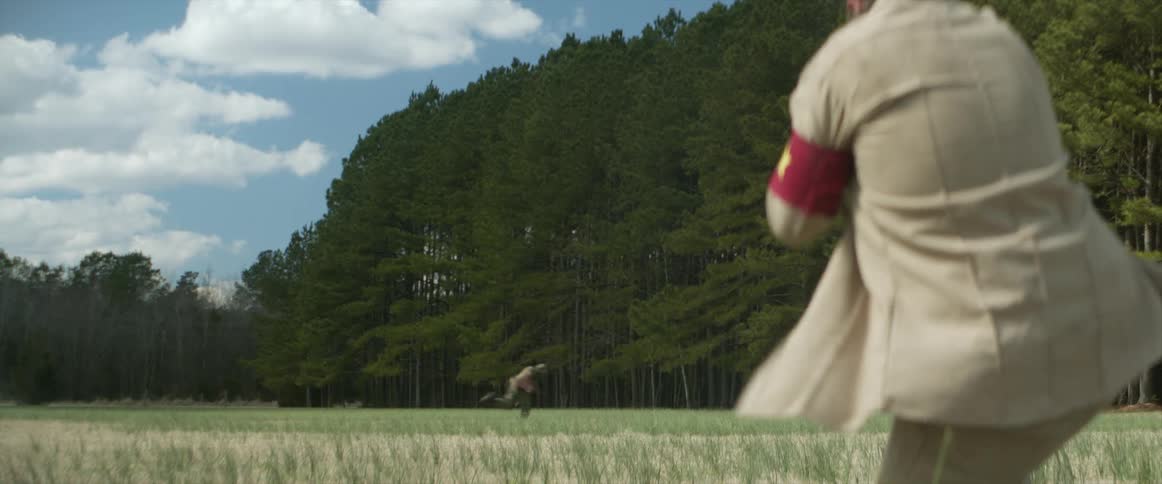 A man with a armband and a gun shooting at another man running with trees the sky and clouds in the background