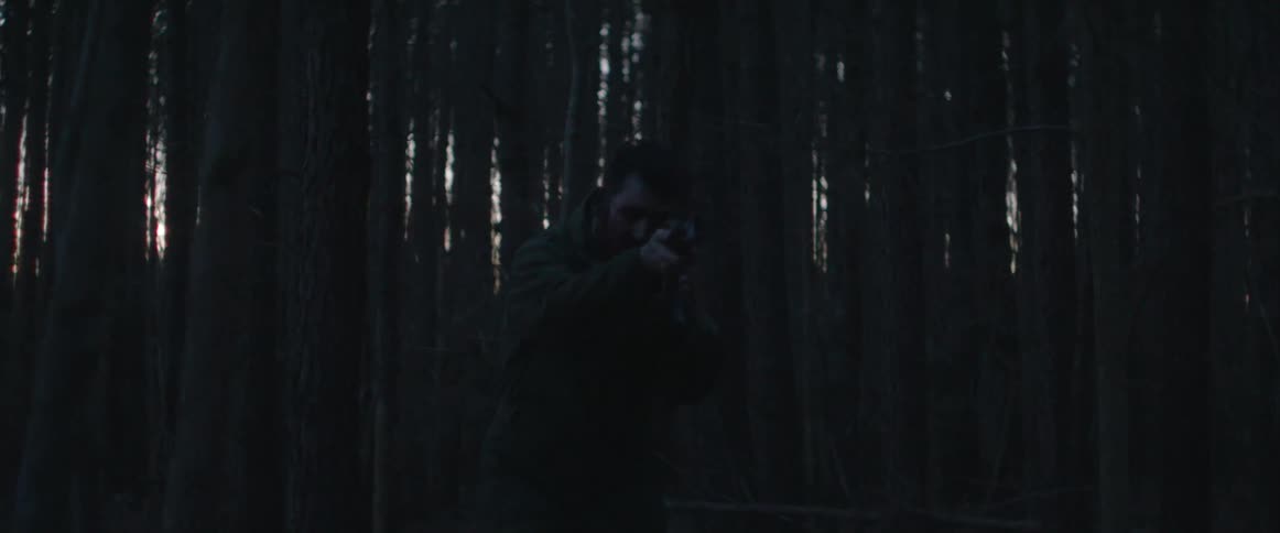 A man walks slowly through a dark forest while aiming a gun directly in front of him