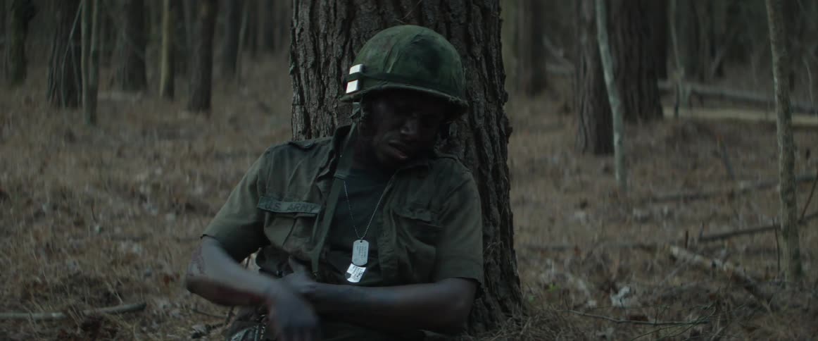A wounded soldier slides to a sitting position with his back against a tree before removing a cigarette from his pocket and placing it in his mouth