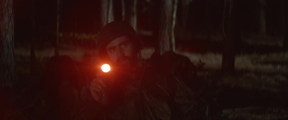 A man sits outside in the dark holding a flashlight and writing in a book and then he stands up and looks off in to the distance and raises a gun