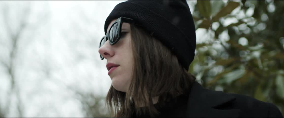 A woman with a hat and sunglasses standing with trees the sky and clouds in the background