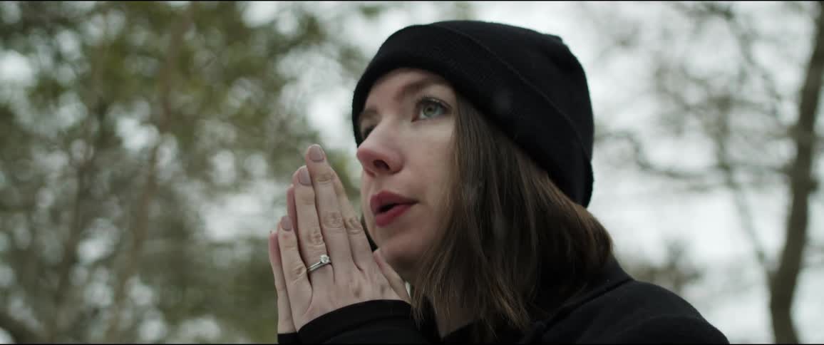 A woman with a jacket and a hat standing outside while its snowing with trees the sky and clouds in the background