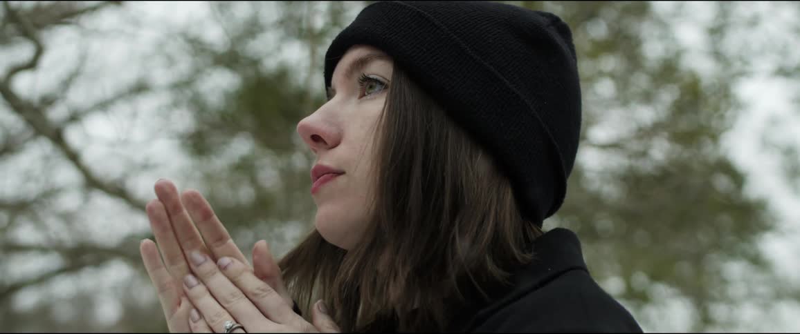 Woman wearing a black knit hat and a black coat is rubbing her hands together as snow falls and trees are behind her