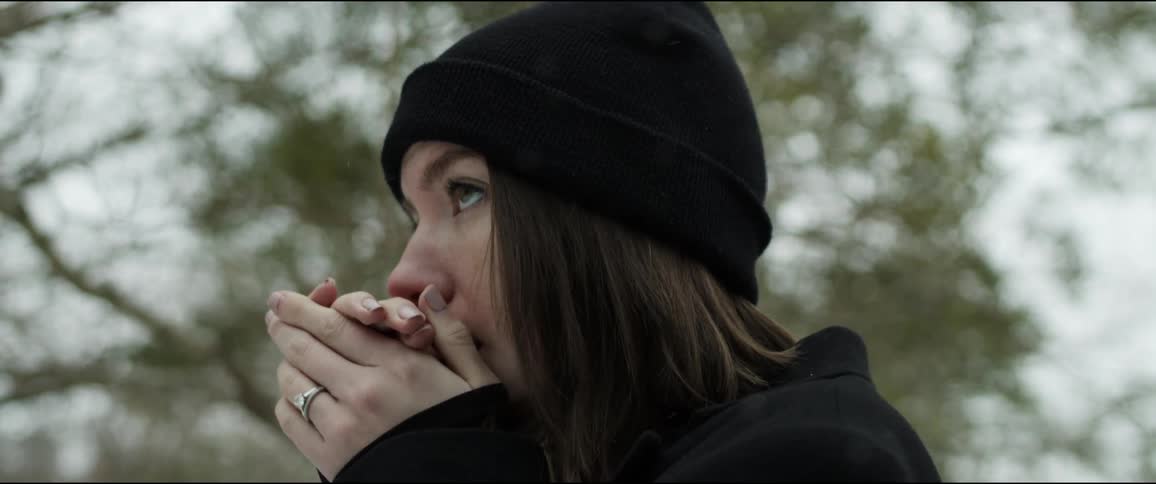 Young woman wearing a black sweater and beanie warms her hands with her breath standing outdoors in the winter