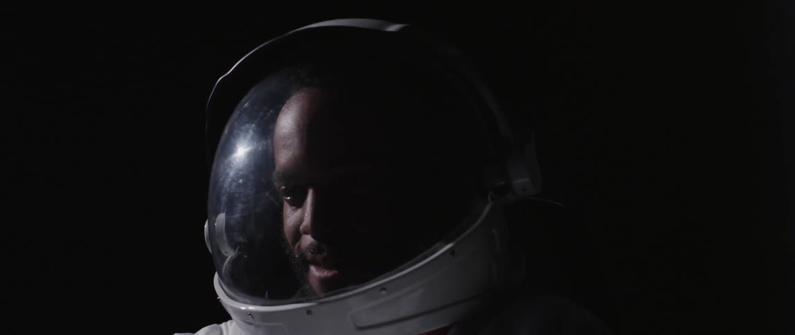 A man in an astronaut helmet looks around and reaches upward