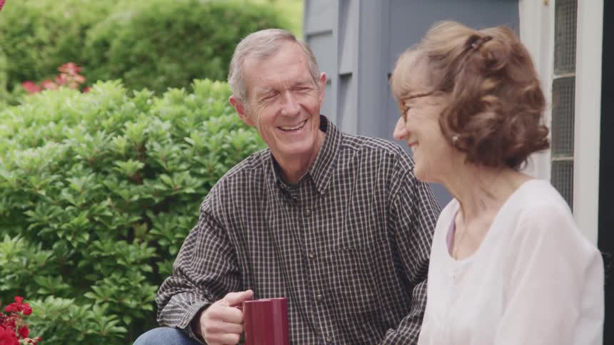 An elderly man and woman sit outside their home and laugh together