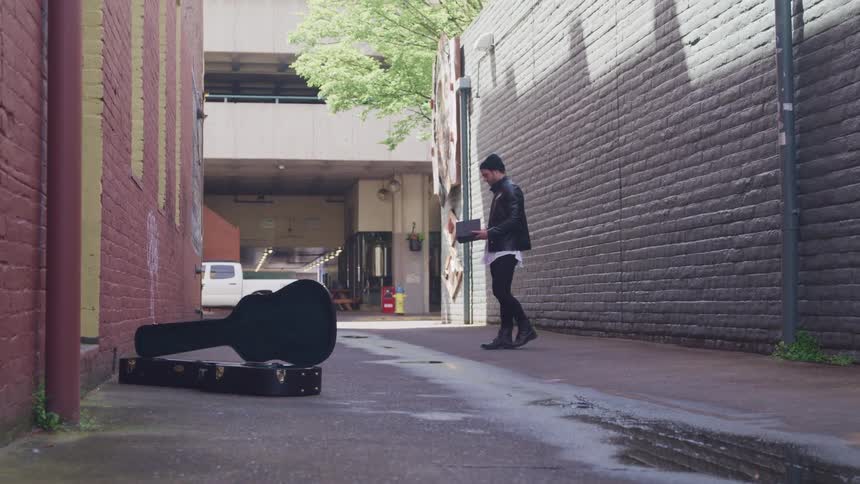 A young musician in an alley with a guitar off to the side is holding a journal and then throwing with anger