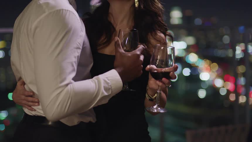 Young couple wearing formal outfits slowly dancing with wine glasses on their hands in front of the city at night