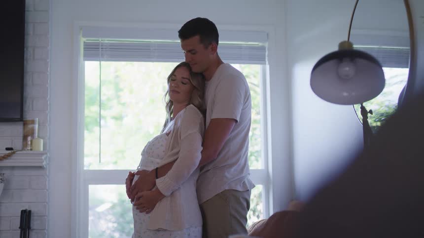 A man and a woman standing in a room hugging with a wall and window in the background
