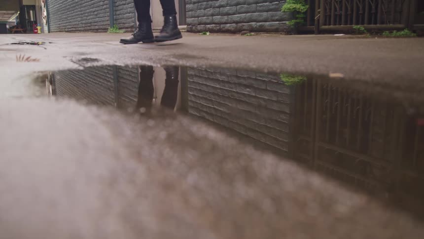 A person with boots walking by a puddle with a wall and a door in the background