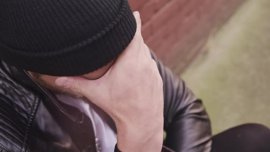 A depressed man sits outdoors with his back against a brick wall as he massages the bridge of his nose with his fingers
