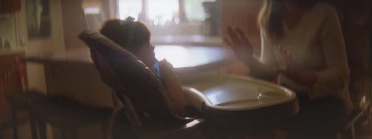 A mother in the kitchen coaxes her baby sitting in a highchair