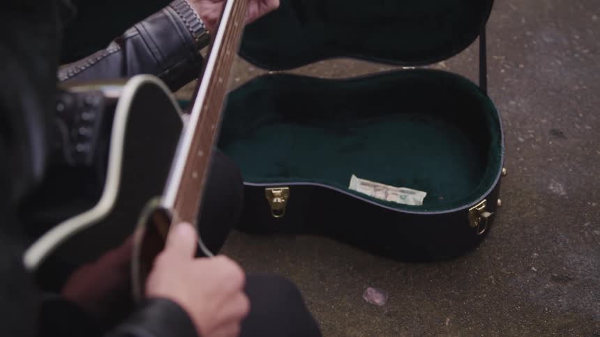 A person with a jacket playing an acoustic guitar while another person puts money in the guitar case