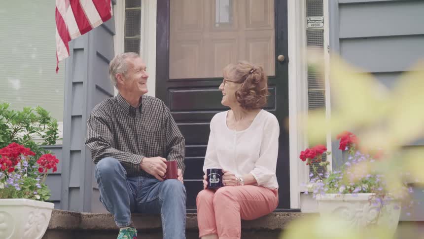 An elderly couple smiling and drinking coffee on a front porch on a summer day
