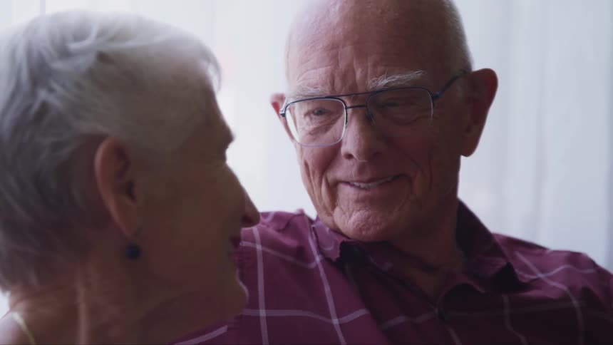 An elderly man and woman sit at home and smile at one another as they look in to each others eyes