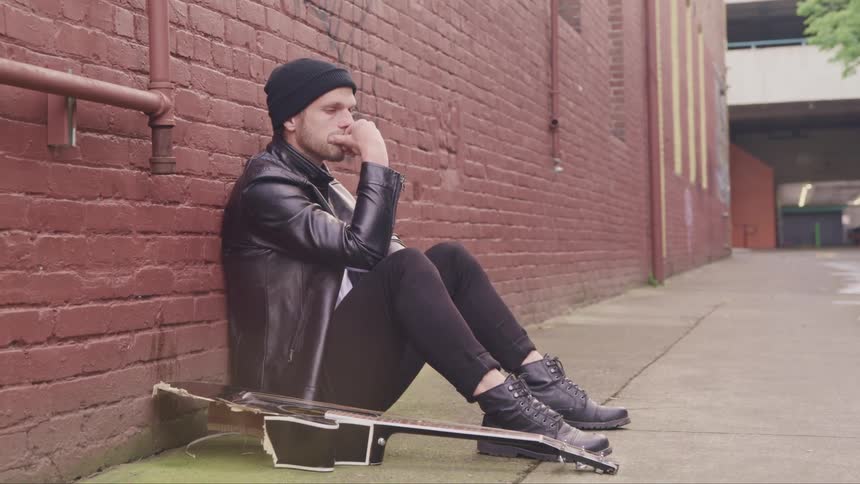 A man sitting outside up against a building next to a broken guitar looks around sadly