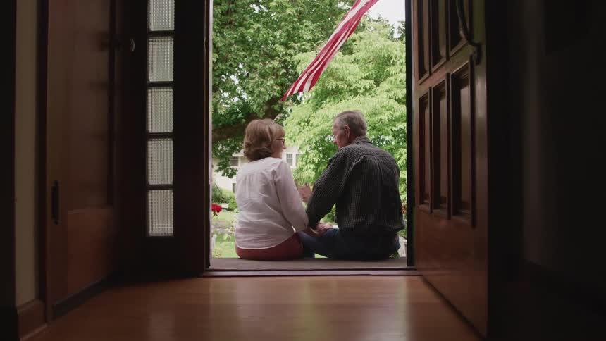 Happy elderly couple sitting on the steps at the entrance of a house looking at each other under the american flag