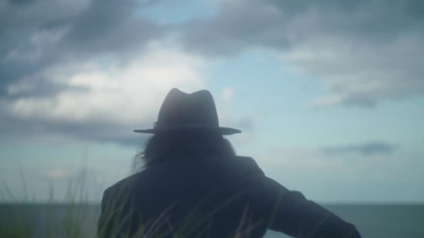 Long haired man wearing a hat plays guitar looking at the sea