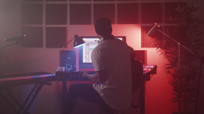 Young man sitting in front of a computer plays acoustic guitar in a studio