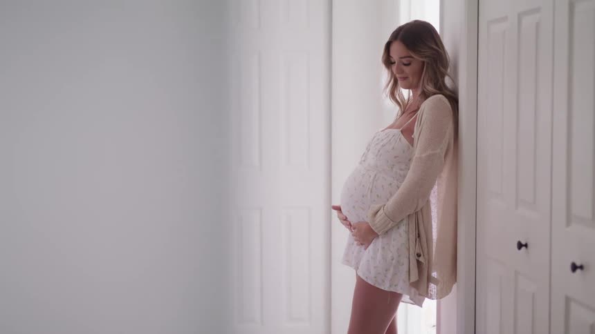A pregnant woman caresses her stomach while standing in a corridor in a house during the day