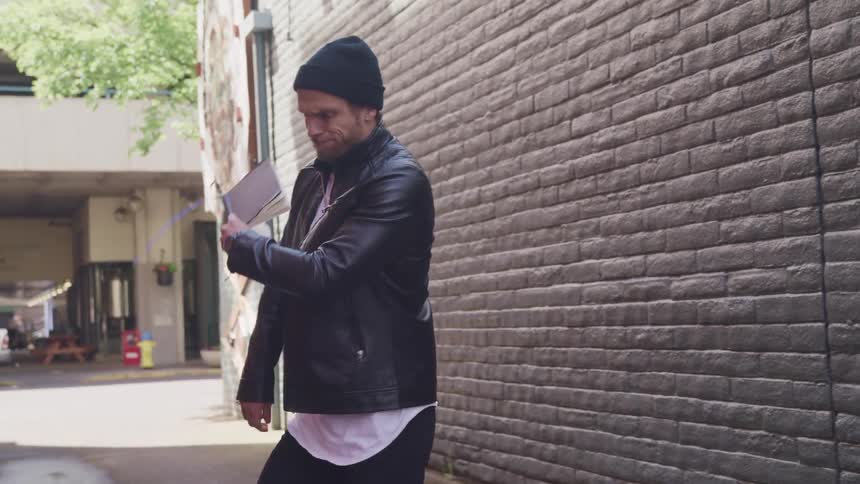 A young man in an alley taps a pencil on a book and then turns around and throws the book against a brick wall
