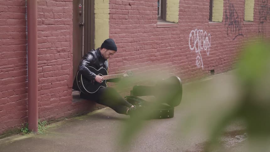 A man is playing guitar and sitting in an alley