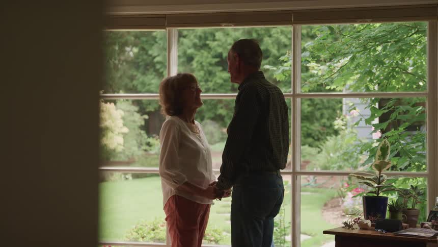 A husband and wife holding hands and looking at each other in front of a window looking out into a beautiful backyard