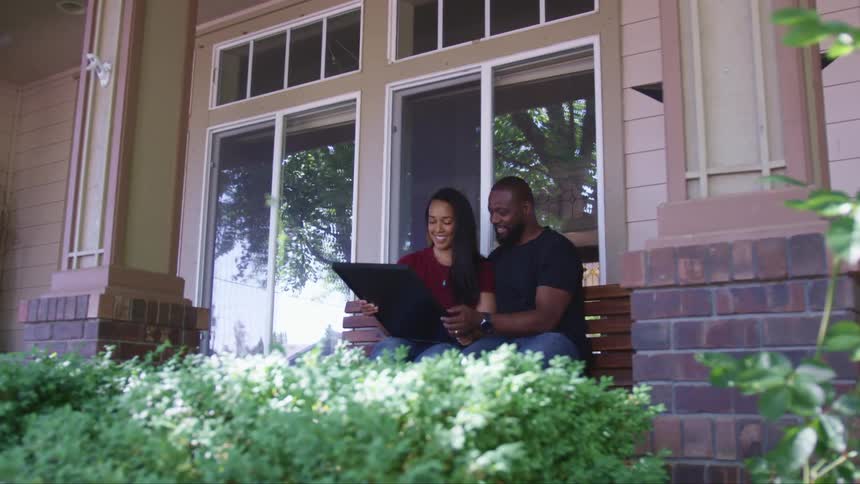 A man and a woman sitting on a bench on a porch in front of a house looking at a picture
