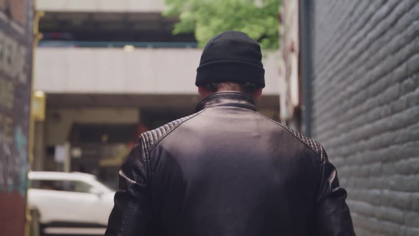A man in a leather jacket walking in an alley in a city
