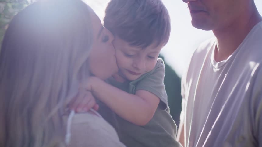A little boy hugs his mother who holds him in her arms and she kisses his face and his dad leans down and kisses him on the head