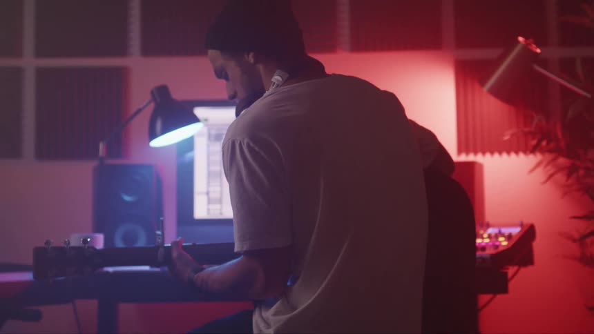 Young man sits in front of a computer with an acoustic guitar inside a studio and starts recording