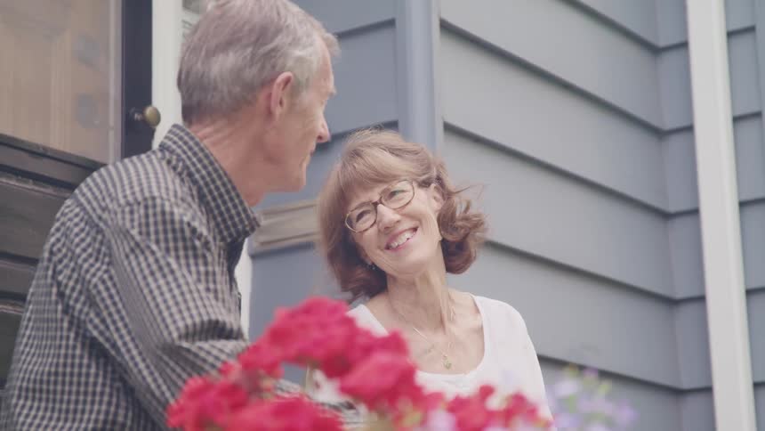 An elderly couple laugh and talk with one another while sitting outside the door of a house during the day