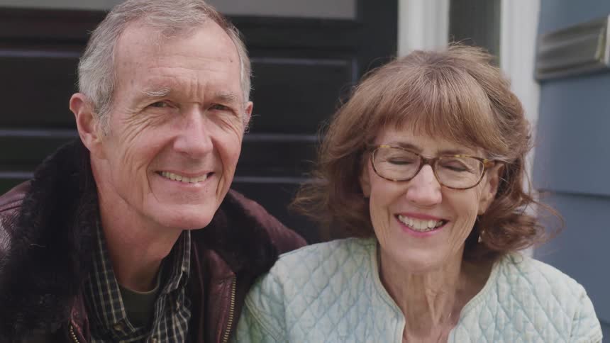 Happy elderly couple sitting in the setps at the entrance of a house looking at the camera