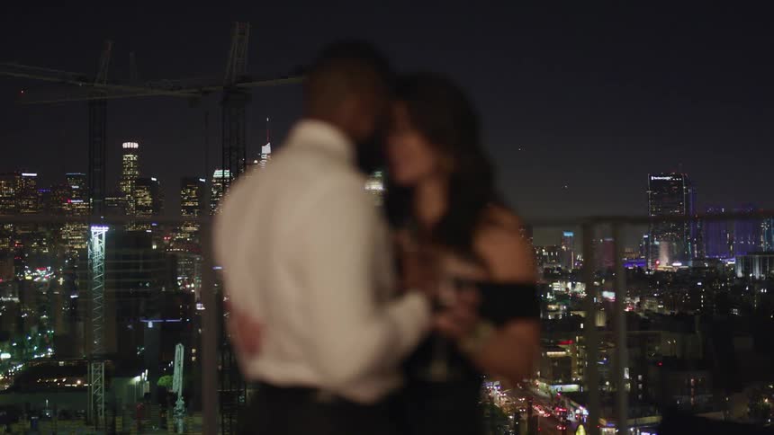 A man and woman slow dance on a rooftop with construction cranes and the city skyline behind them