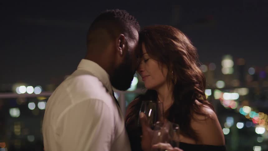 A man and a woman holding wine glasses and standing on a rooftop with their foreheads touching with the city skyline in the background