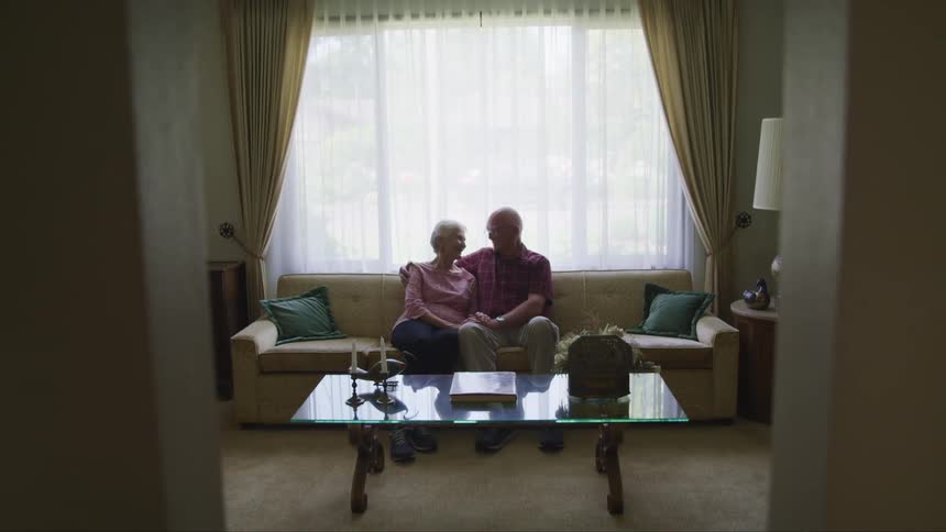 A man has his arm around his wife as they sit on the couch and smile at each other