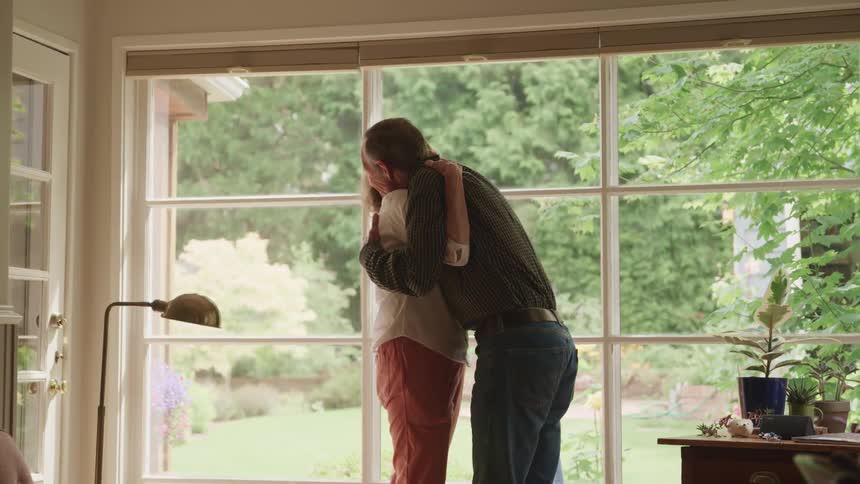 An elderly couple hugging in front of a living room window at home