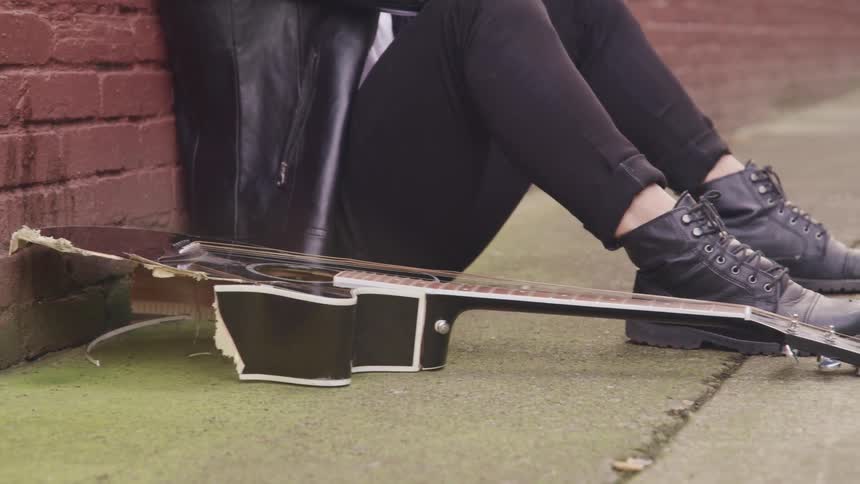 A person sits on the floor of an alleyway besides a broken acoustig guitar